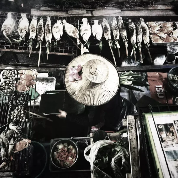 A Thai lady in a traditional straw hat grilling and selling fresh fish skewers from her boat at a floating market, an authentic cultural and food explorer experience.